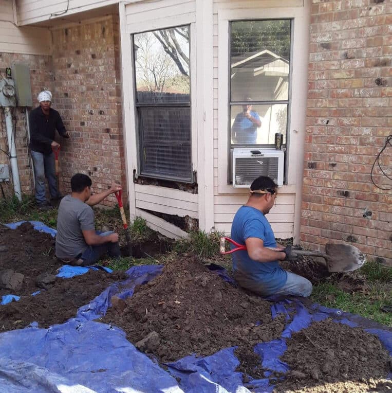 three foundation repair technicians digging holes along the outside of a home's foundation