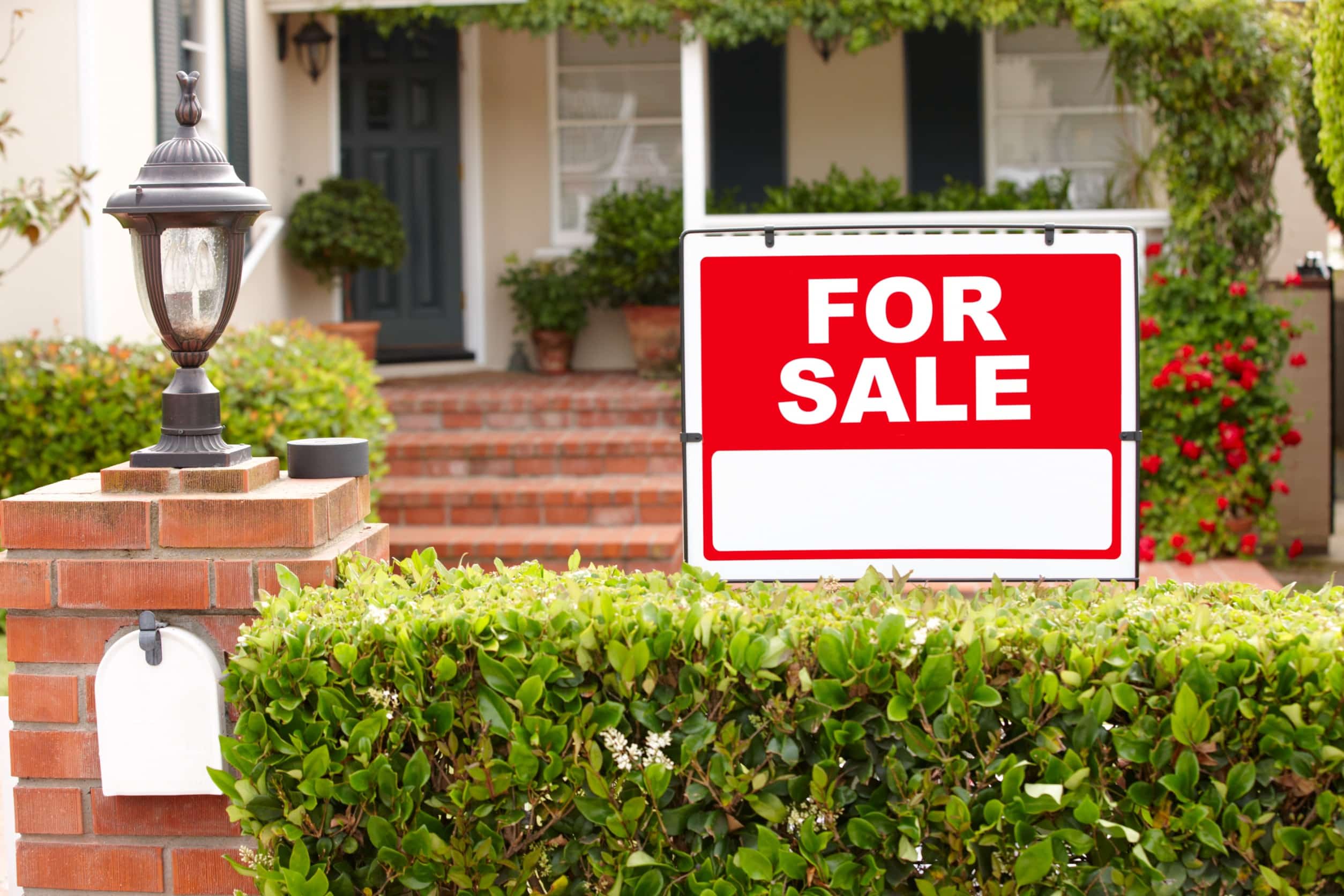 a red and white sign reading, "for sale" in the bushes outside a white home with red brick stairs and blue shutter
