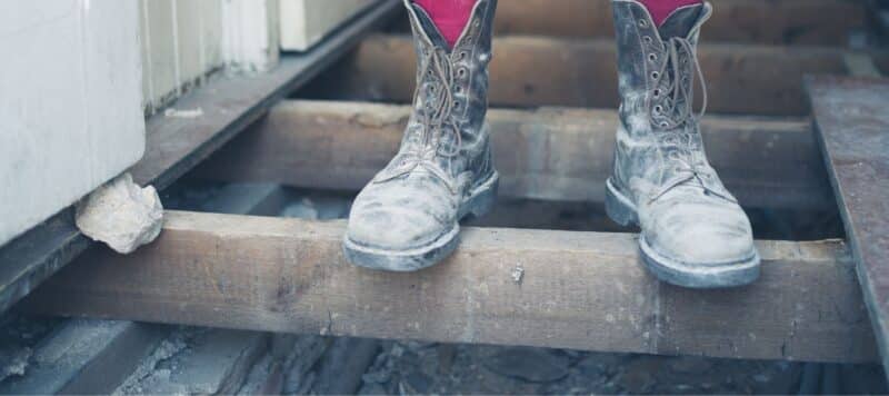 Close-up of dusty, worn work boots standing on a wooden beam at a construction site. The boots are grey with visible signs of wear and dirt, highlighting their frequent use in a rugged environment. The setting appears to be an incomplete structure with exposed wooden beams underfoot