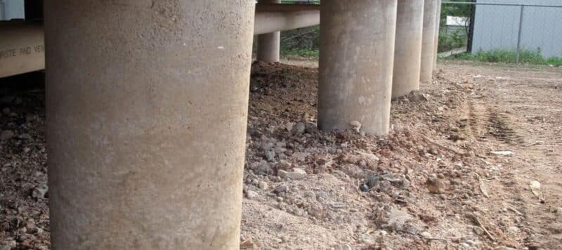 A close-up view of several thick concrete pillars supporting a structure above. The ground is dry and scattered with debris and small rocks, indicating recent construction or excavation work in the area. The focus on the pillars emphasizes their robust and solid construction, typical of infrastructure supports