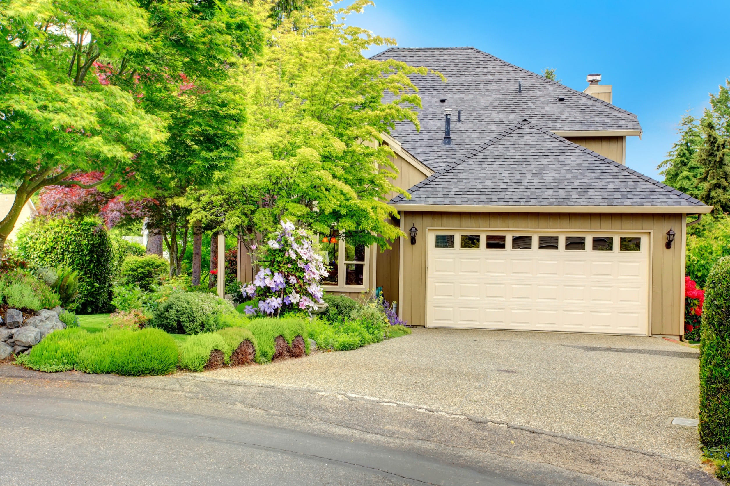 front view of tan home with white garage on a sloping driveway with a large tree in the front yard