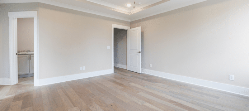 Empty room with light wooden flooring, beige walls, white trim, an open door to an adjacent space, a bathroom door with a visible sink, and a modern tray ceiling with recessed lighting