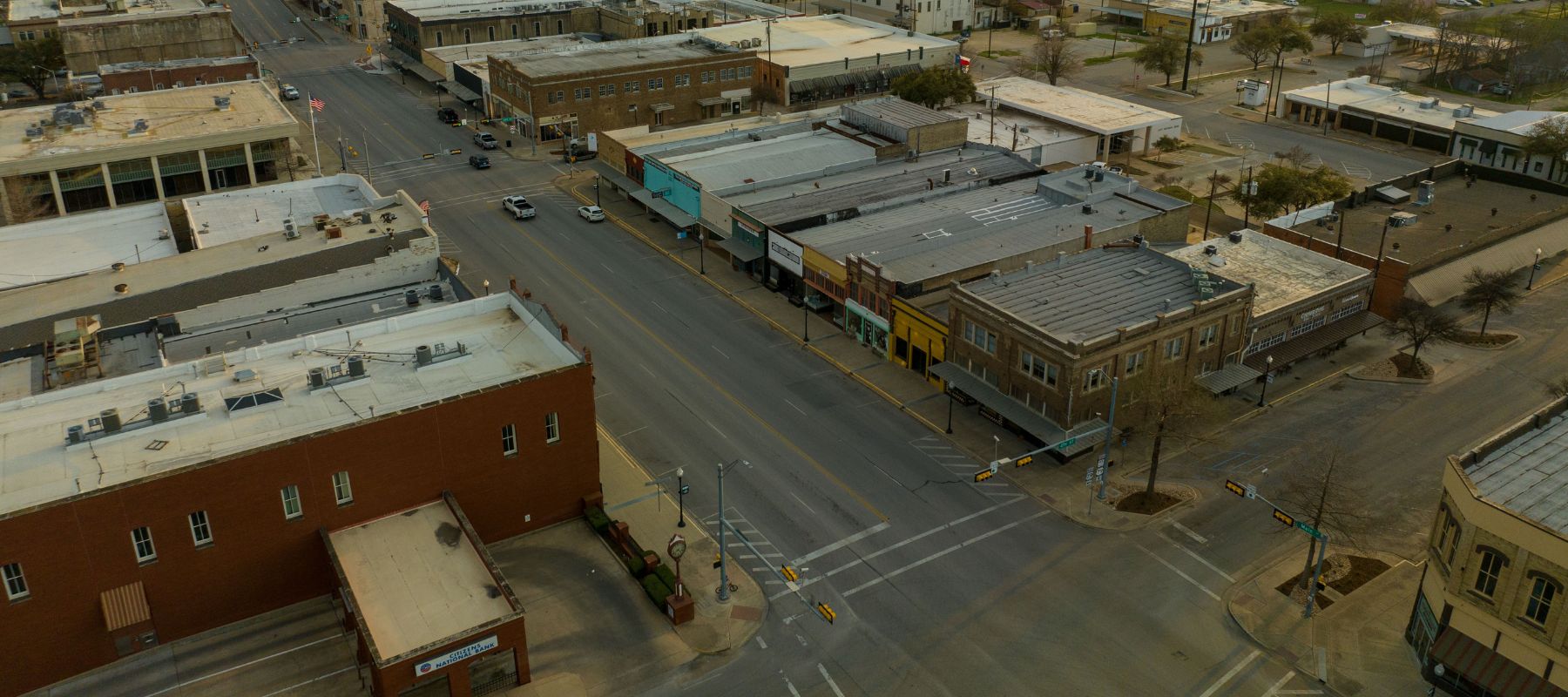An aerial view of a small town's downtown area shows a main street with older buildings, cars parked along the road, and an intersection